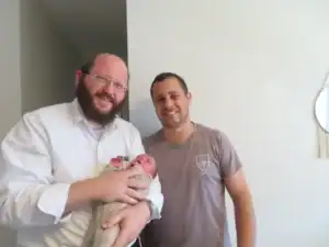 מוהל מקצועי בתל אביב מחזיק תינוק לאחר טקס ברית מילה, לצד האב המחייך